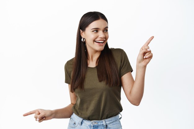 Young millennial girl with joyful smile going on shopping, facing choice, pointing sideways and picking product in store, having variants