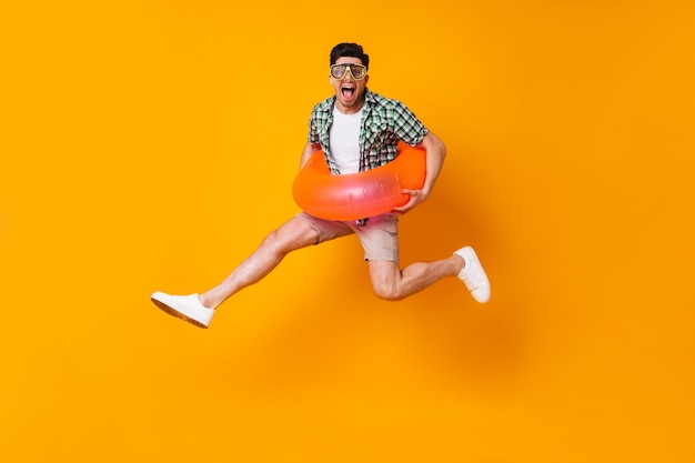 Young mischievous man in summer outfit and diving mask jumping with inflatable circle on orange space.