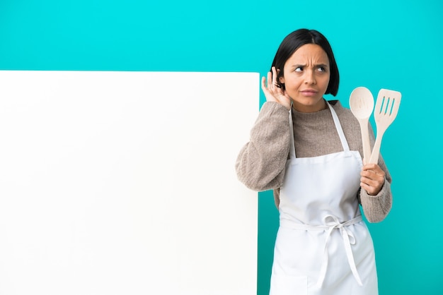 Young mixed race cook woman with a big placard isolated on blue background listening to something by putting hand on the ear