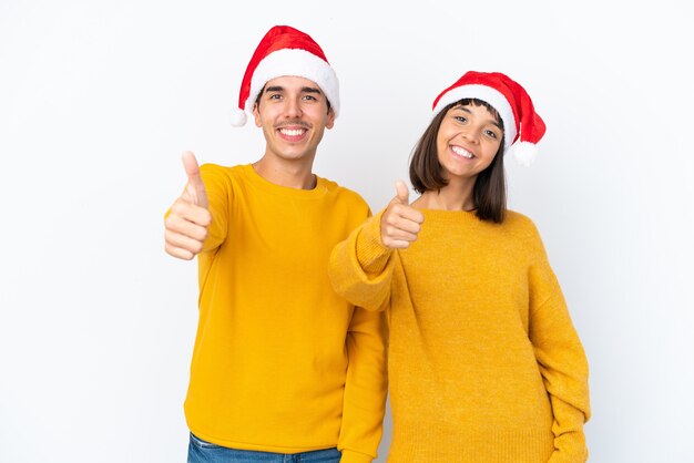 Молодая пара смешанной расы празднует рождество изолированно на белом фоне, показывая большой палец вверх, потому что произошло что-то хорошее