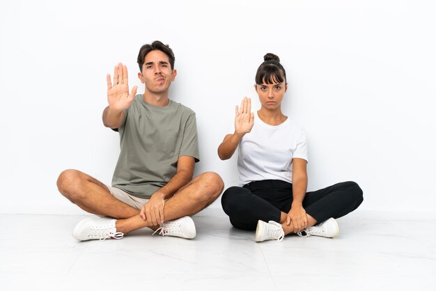 Young mixed race couple sitting on the floor isolated on white background making stop gesture denying a situation that thinks wrong