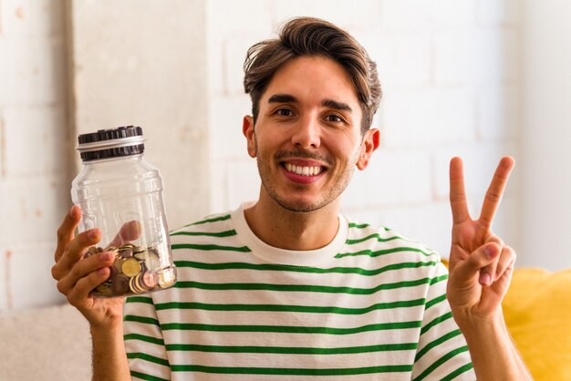 Young mixed race man holding piggy bank in his living room joyful and carefree showing a peace symbol with fingers.