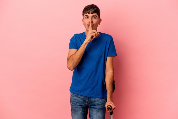 Young mixed race man with crutches isolated on pink background keeping a secret or asking for silence.