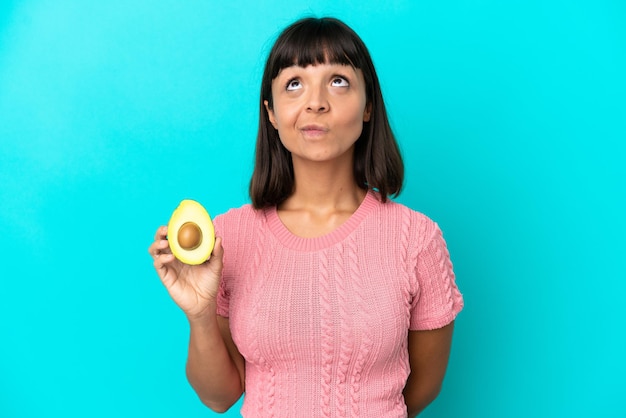 Молодая женщина смешанной расы держит авокадо на синем фоне и смотрит вверх