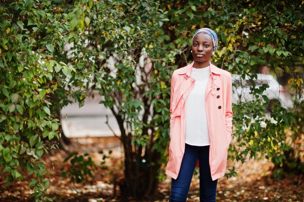 Young modern fashionable attractive tall and slim african muslim woman in hijab or turban head scarf and pink coat posed at park