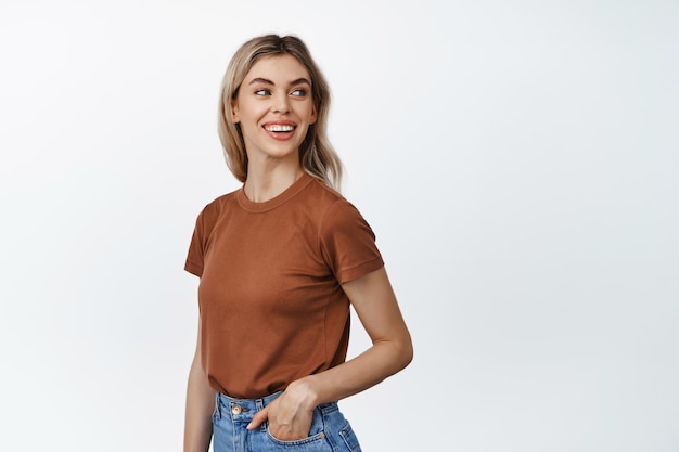 Young modern girl with blond hair looking behind her shoulder to the right side and smiling friendly standing against white background