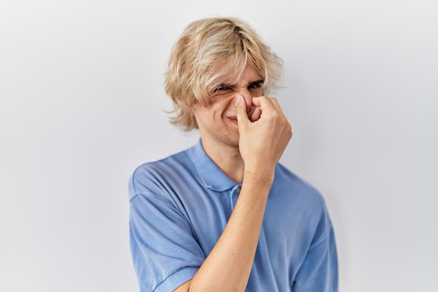 Young modern man standing over isolated background smelling something stinky and disgusting intolerable smell holding breath with fingers on nose bad smell