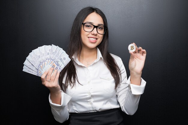 Young modern successful business woman in glasses holding a bitcoin and cash money isolated on black wall