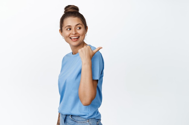 指を右に向けて、幸せな笑顔で彼女の肩の後ろを見て、青いTシャツとジーンズを着て、白い背景の上に立っている若い現代女性