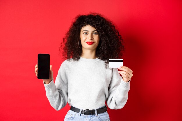 곱슬머리를 한 젊은 현대 여성이 플라스틱 신용 카드와 휴대 전화 화면을 보여주고 온라인 쇼핑 앱을 보여주고 빨간색 배경에 서 있습니다.