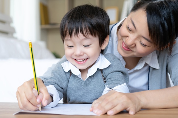 Young mom caught son's hand holding a pencil to measles write down on white paper, preschool at home