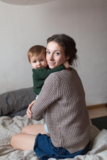 침대 컨셉의 홈 컴포트 라이프스타일에서 노는 니트 스웨터를 입은 젊은 엄마와 아기