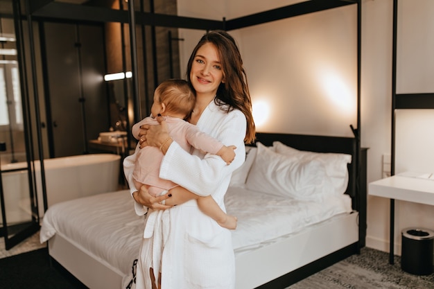 Young mother in bathrobe holding baby in diaper and looking at camera with smile against white bed.