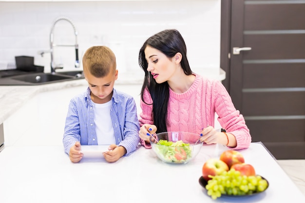 若い母親が息子が台所で電話ゲームで遊んでいる間にサラダを調理する