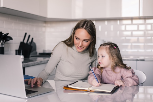 Молодая мать помогает дочери с домашним заданием за столом в столовой