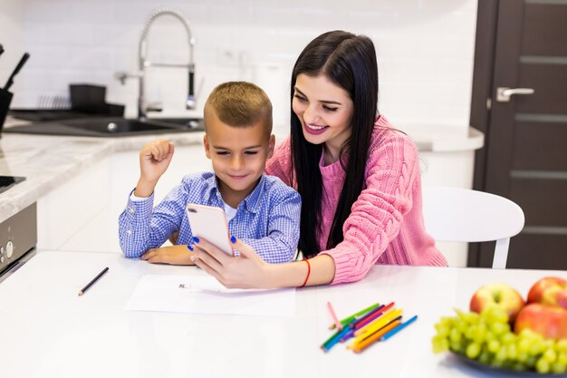 Young mother holding phone and show screen with win gesture in the kitchen