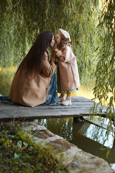 Молодая мать в пальтоме объявляет свою маленькую дочку, сидящую на мосте у озера и филиалов
