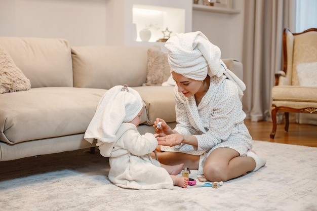 Young mother and little baby girl wearing dressing gowns with hair wrapped in towels Woman and girl sitting on a floor near sofa Woman painting a nails of her daughter