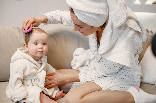 Young mother and little baby girl wearing dressing gowns with hair wrapped in towels Woman and girl sitting on a sofa Woman combing a hair of her daughter