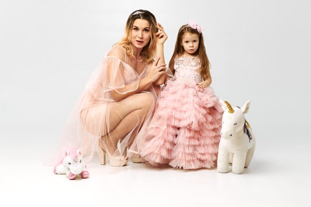 Молодая мать сидит на полу вместе с милой очаровательной маленькой дочкой в причудливых розовых одеждах, играя с игрушками