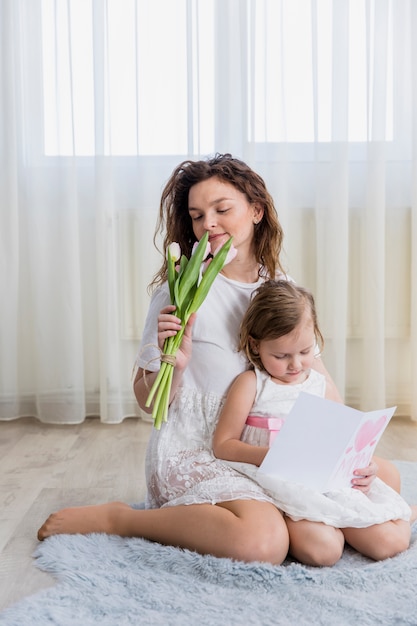 Young mother smelling flowers while little daughter reading greeting card at home