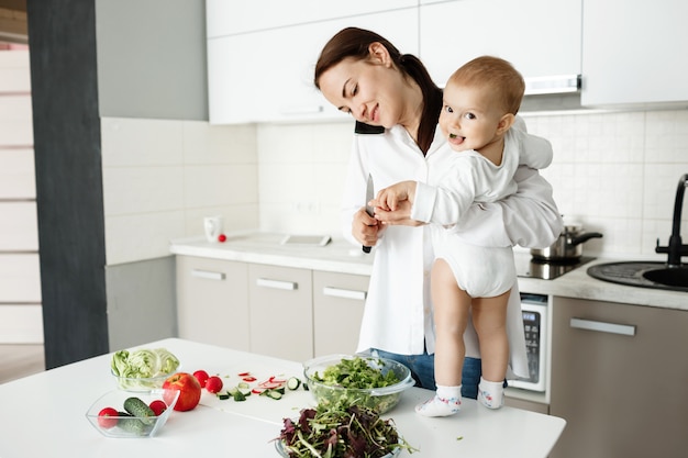 Young mother taking care of little child, talking on phone and cooking at same time