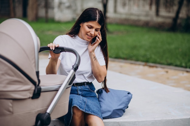 Young mother walking in park with baby stroller and talking on phone