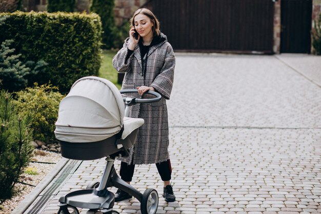 Young mother walking with baby carriage in park and talking on the phone