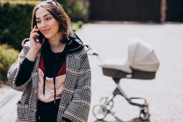 Young mother walking with baby carriage in park and talking on the phone