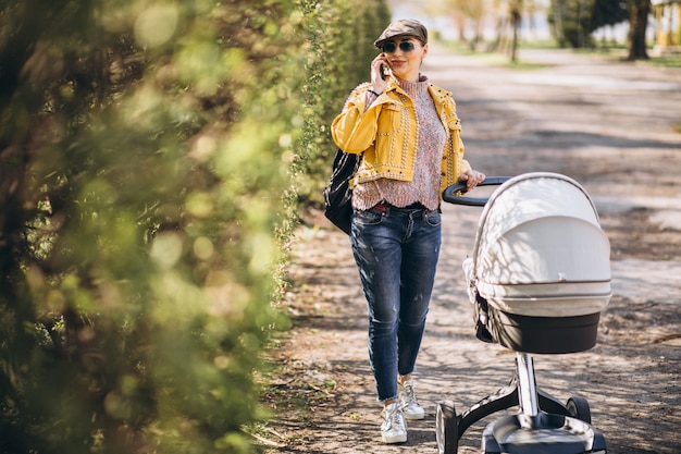 Young mother walking with baby carriage in park and using phone