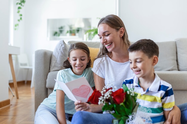 Young mother with a bouquet of roses laughs, hugging her son, and ?heerful girl with a card and roses congratulates mom during holiday celebration at home. mothers day