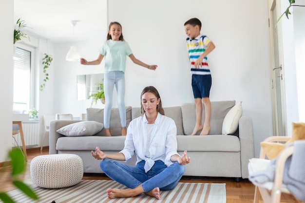 Young mother with closed eyes meditating in lotus pose on floor trying to save inner harmony while excited children jumping on sofa and screaming in light spacious living roo