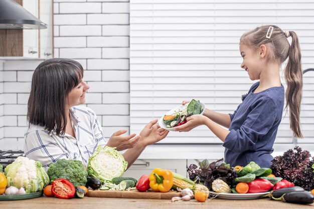 Young mother with her daughter with many vegetables on the background of the interior of a modern light kitchen.