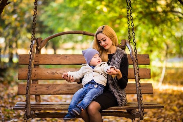 Молодая мать с маленьким сыном отдыхает на деревянной скамейке в осеннем золотом парке