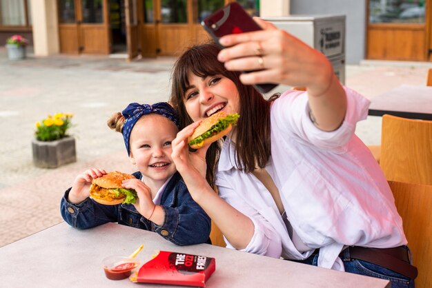 Young mother with little girl eating a hamburger take selfie on the street cafe