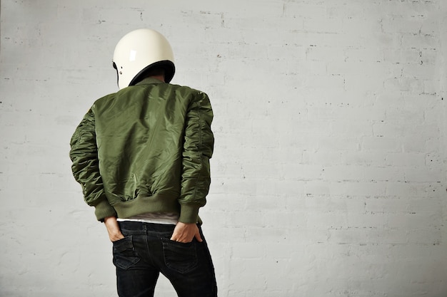 무료 사진 흰색 벽과 그의 청바지 뒷 주머니에 그의 손으로 뒤에서 흰색 헬멧과 녹색 재킷 초상화에 젊은 오토바이.