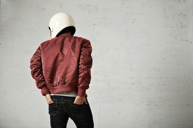 Young motorcyclist in white helmet and red nylon jacket shot from the back with his hands in the back pockets of his jeans in a studio with white walls.