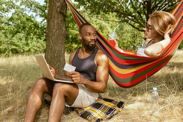晴れた夏の日の牧草地で屋外で若い多民族の国際的なロマンチックなカップル。一緒にピクニックをしているアフリカ系アメリカ人の男性と白人女性。関係の概念、夏。