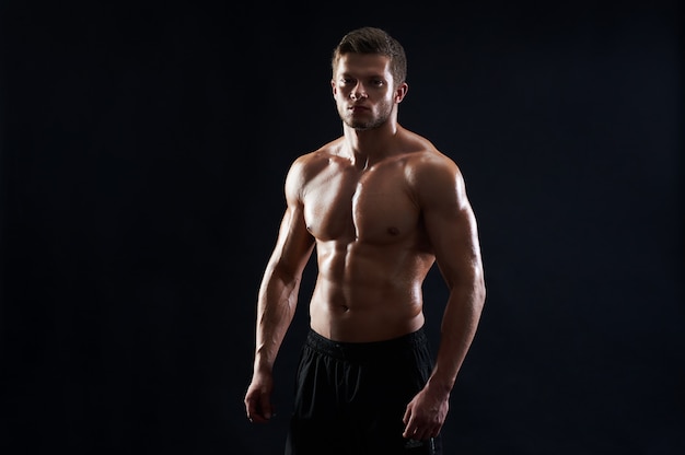 Молодой мускулистый спортсмен позирует без рубашки на черном фоне