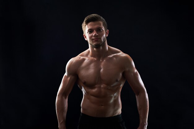 Молодой мускулистый спортсмен позирует без рубашки на черном фоне