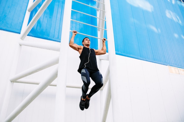 young muscular man doing pull ups, listening to music in headphones during workout