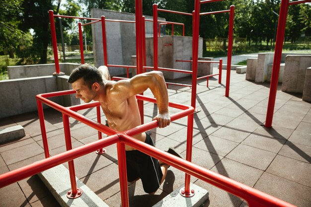 Молодой мускулистый кавказский мужчина без рубашки делает подтягивания на турнике на детской площадке в солнечный летний день. Тренирует верхнюю часть тела на открытом воздухе. Понятие спорта, тренировки, здорового образа жизни, благополучия.