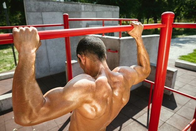 Foto gratuita giovane uomo caucasico senza camicia muscolare che fa pull-up sulla barra orizzontale al parco giochi nella soleggiata giornata estiva. allenare la parte superiore del corpo all'aperto. concetto di sport, allenamento, stile di vita sano, benessere.