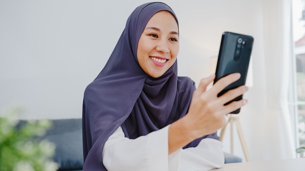 Young muslim businesswoman using smart phone talk to friend by videochat brainstorm online meeting while remotely work from home at living room.