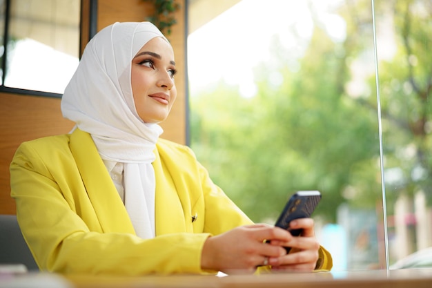 カフェで彼女のスマートフォンを使用してヒジャーブの若いイスラム教徒の女性