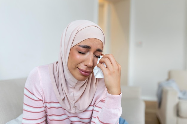 Молодая мусульманка, сидящая в комнате, чувствует боль от жизненных проблем Плачущая мусульманка, держащая болезненную руку, страдающую от насилия мужа