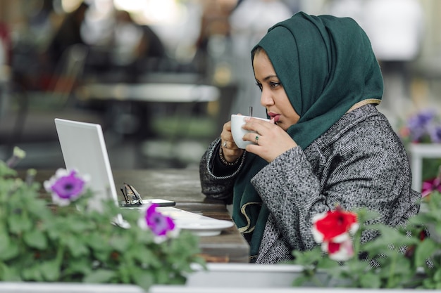 通りのカフェに座って、ラップトップで見ている若いイスラム教徒の女性
