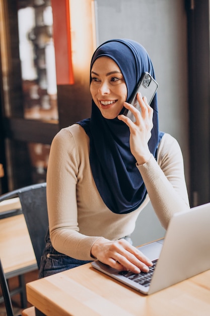 전화를 사용하고 카페에서 컴퓨터에서 작업하는 젊은 무슬림 여성