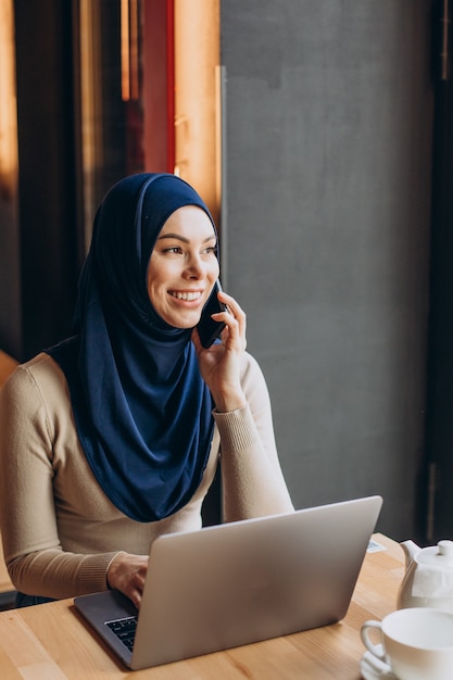 電話を使い、カフェでコンピューターを操作する若いイスラム教徒の女性