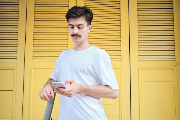 Молодой усатый мужчина в белой футболке задумчиво пользуется мобильным телефоном на желтом фоне на городской улице